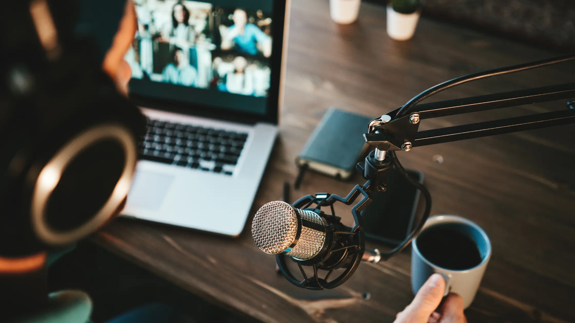 A photo looking over someone's shoulder as they podcast, with microphone, coffee, and laptop on their desk.